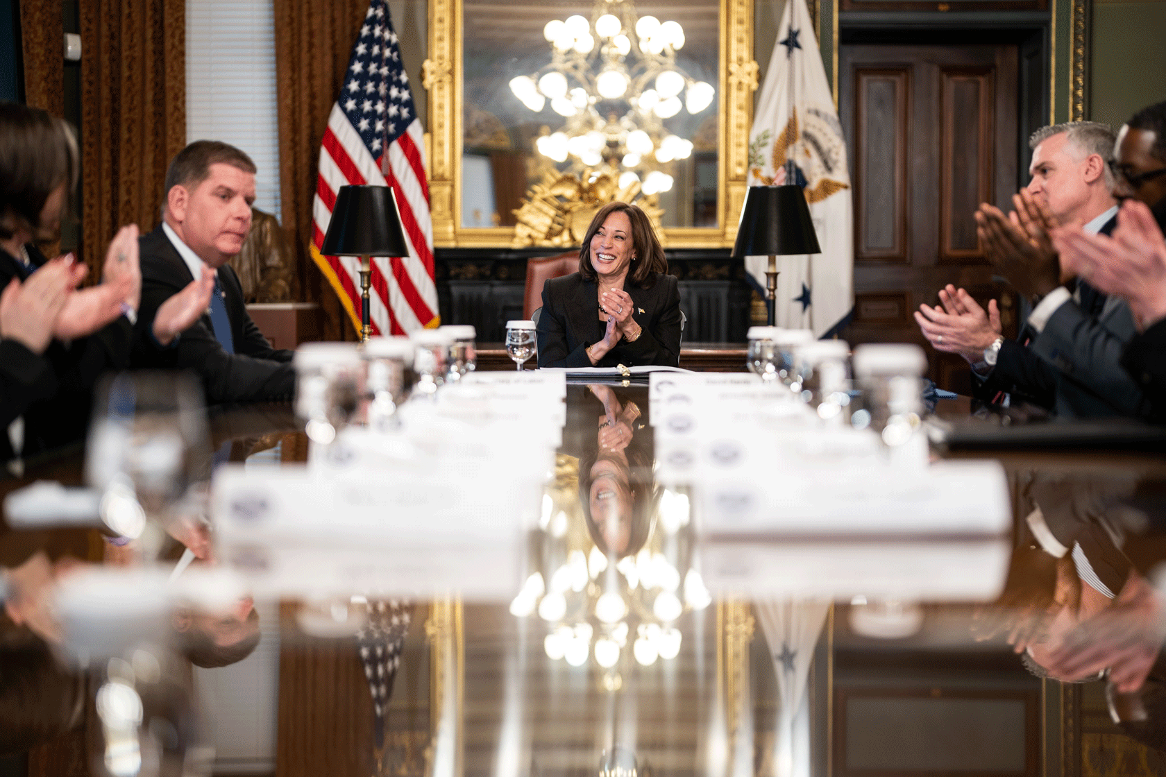 Vice President Kamala Harris is seen smiling and clapping her hands at the end of a long table filled with people.