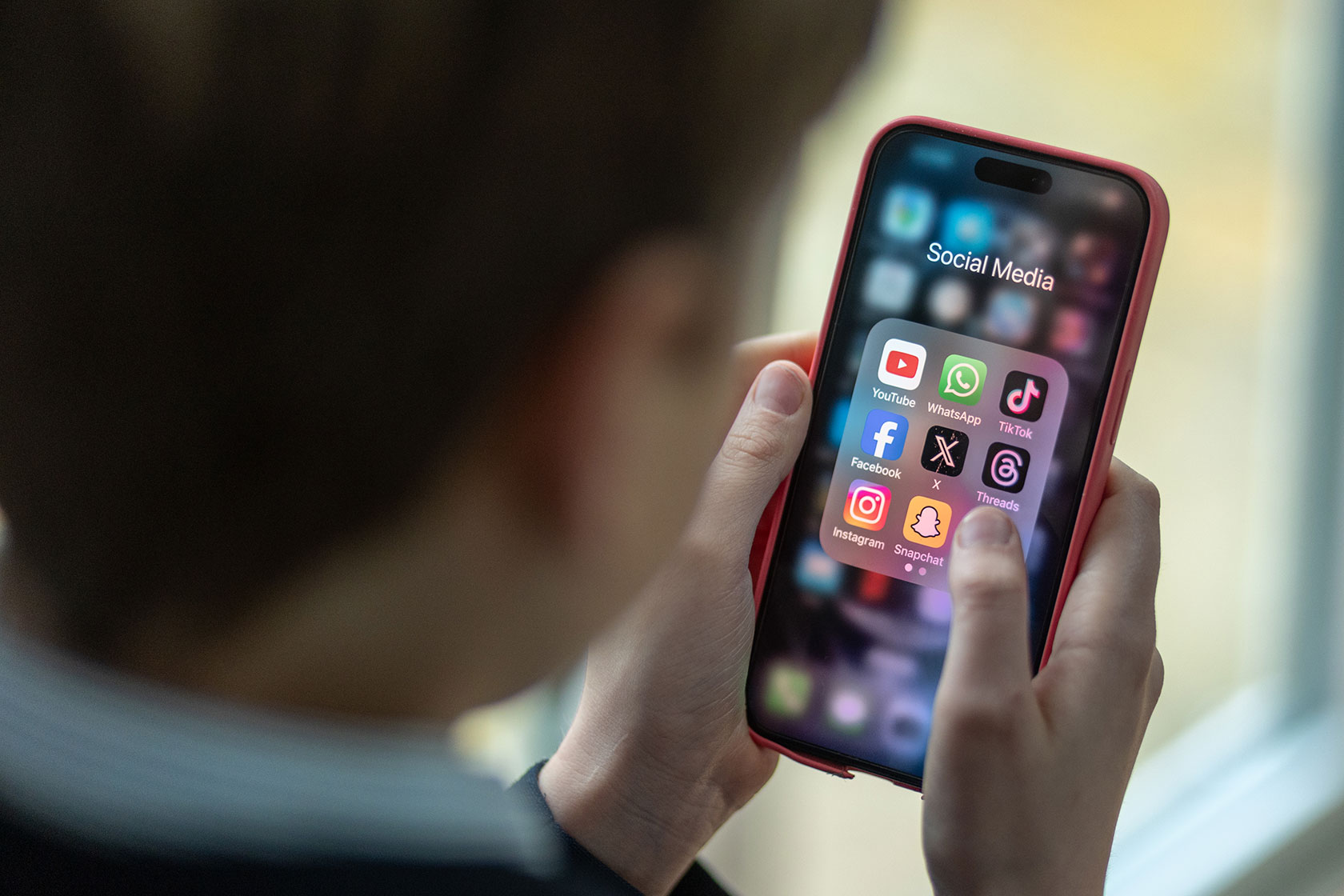 Photo shows a person's hands cradling a phone displaying the Social Media section of the home page, with app icons for YouTube, WhatsApp, TikTok, Facebook, X, Threads, Instagram, and Snapchat