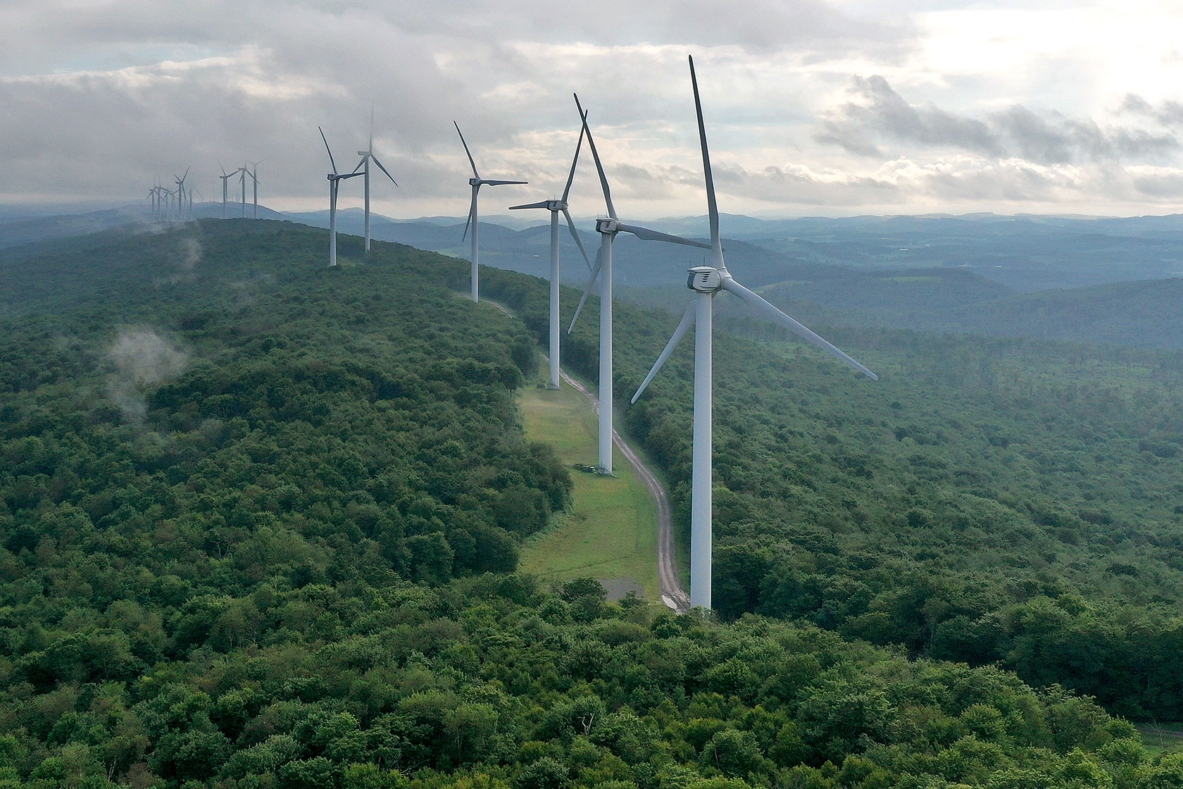 Photo shows a row of white wind turbines standing on the ridge of a green tree-covered mountain, with a cloudy sky in the background